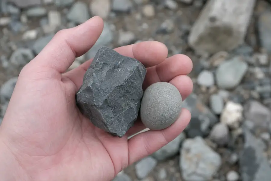 Hand holding angular dark trap rock next to rounded pea stone for comparison