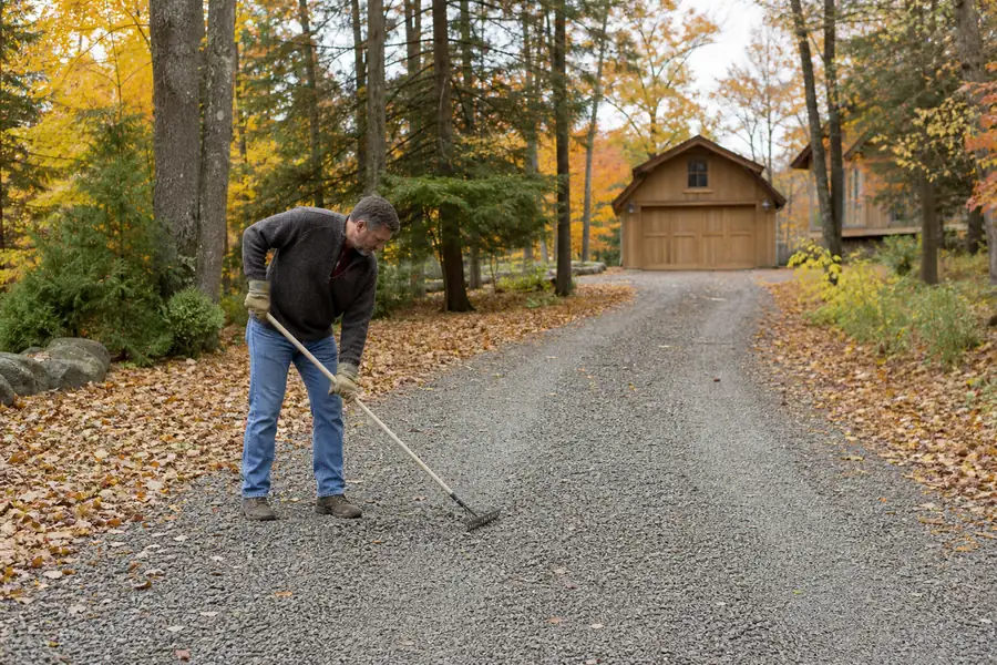 Homeowner raking a gravel driveway on a crisp October day with fall foliage