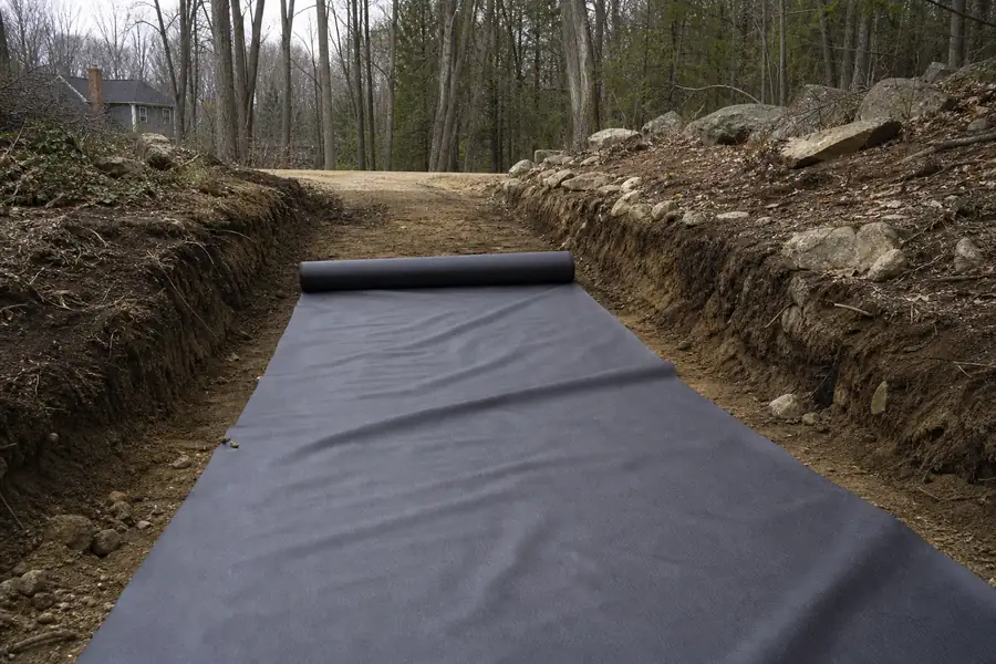 Excavated driveway bed with geotextile fabric being rolled out on a wooded New England lot